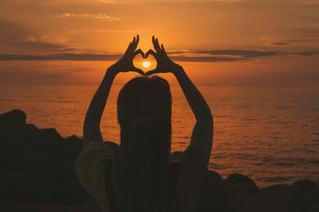 A woman making a heart with her hands in front of a sunset